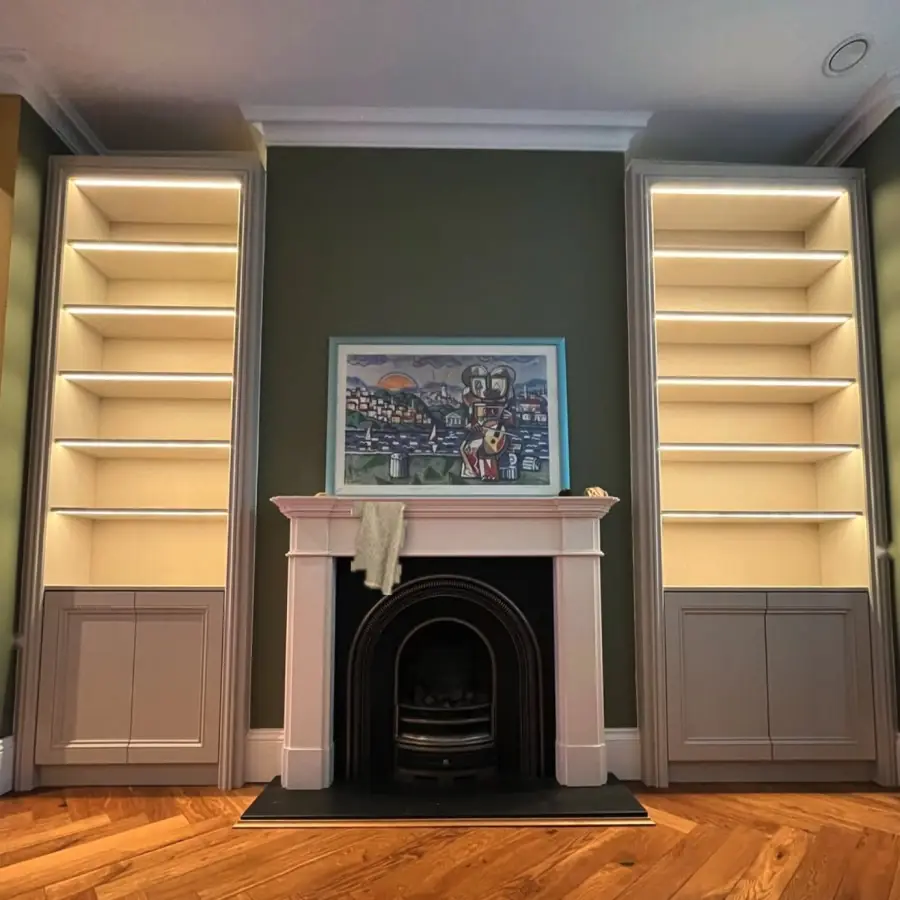 Alcove shelving either side of a fireplace, photographed from low down in yellow light, with bare shelves and a cloth left on the mantel.